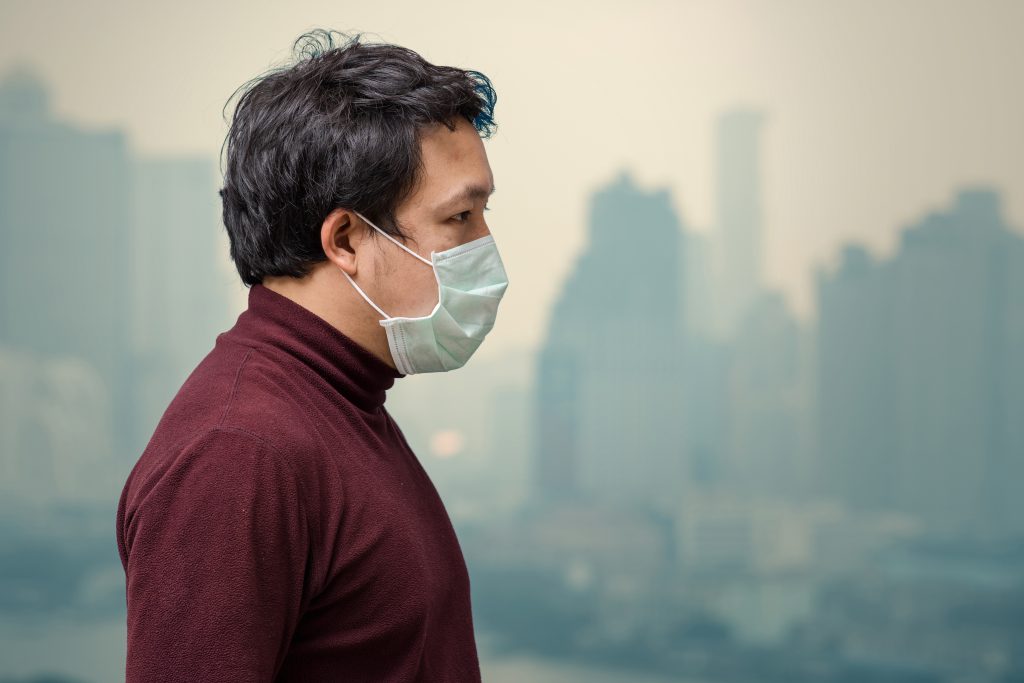 A person wearing a face mask and a maroon sweater stands in profile against a hazy cityscape background, highlighting concerns about environmental factors.