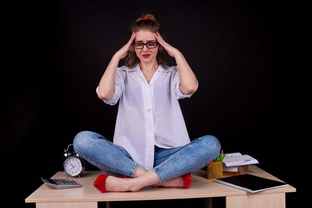 A woman practices mindfulness, sitting cross-legged on a desk with eyes closed, holding her head amidst office supplies, a notebook, and an alarm clock—a calm approach to stress management in a busy workspace.