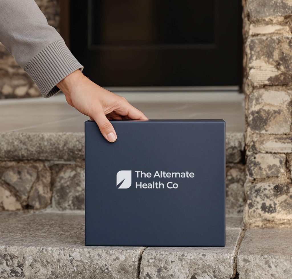 A hand places a navy blue box labeled "The Alternate Health Co" on a stone doorstep in front of a door.
