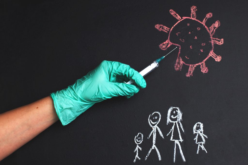 A gloved hand holds a syringe pointed at a chalk drawing of a virus, while a family drawn below highlights the importance of immune support and healthy lifestyle factors on a blackboard background.